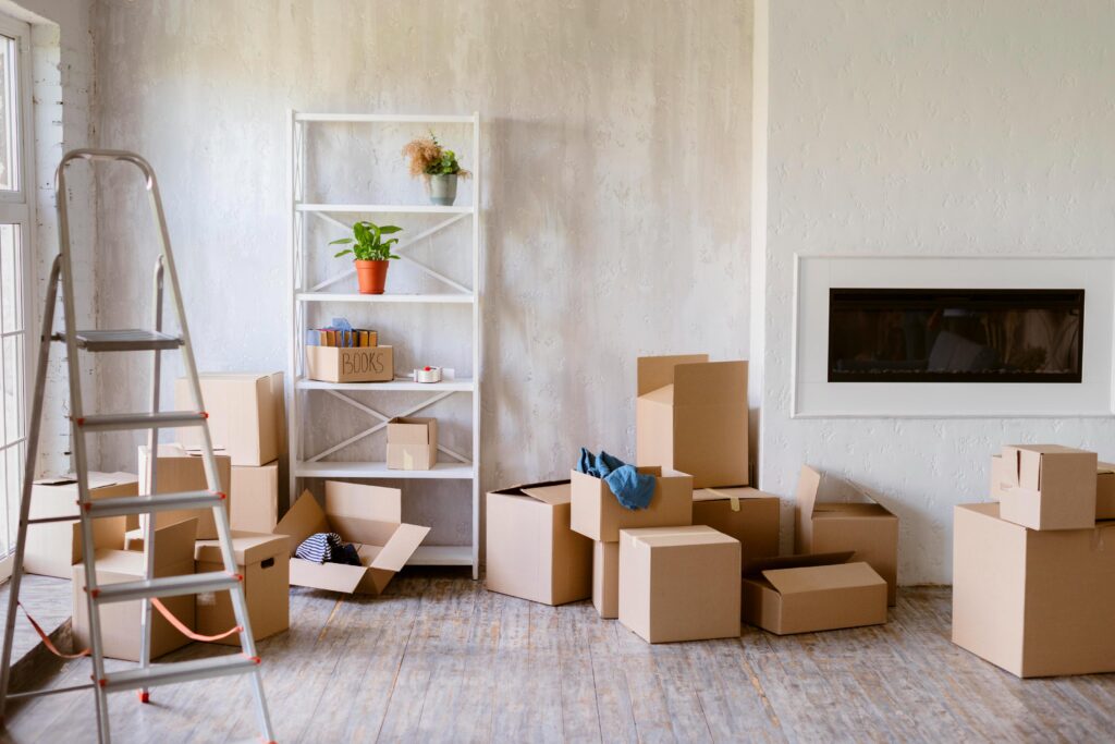 A room with moving boxes on the floor, a fireplace and a shelf unit with boxes for packing and moving.